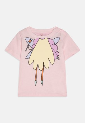 T-shirt in cotone rosa chiaro con una grafica di un personaggio con ali viola, corpo giallo e gambe arancioni, abbinato a scarpe blu e una bacchetta.