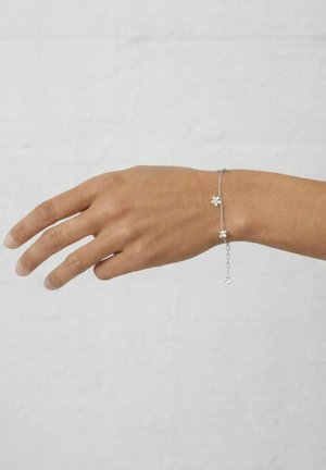 Keskmise nahatooniga käsi kannab delikaatset hõbedast käevõru, millel on väikesed lillekõrvarõngad, ühtlase heleda tausta eest.