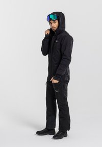 Czarny wodoodporny kombinezon narciarski z kapturem, zamykany na zamek błyskawiczny kieszeniami i regulowanymi mankietami, w komplecie z lustrzanymi goglami i czarnymi butami.