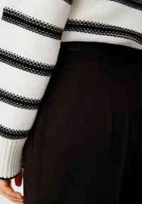 Schwarze Hosen mit einer glatten Textur, kombiniert mit einem weißen Pullover, der dicke schwarze Streifen und einen gerippten Kragen hat.