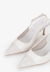Zapatos de tacón slingback de cuero blanco con punta afilada, puntera de charol brillante y un tacón metálico delgado, con un cierre en bucle sutil.