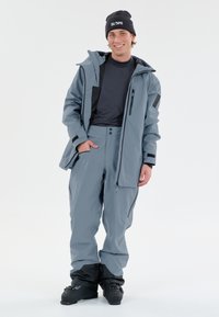 Grå skidjacka och byxor set, med huva, dragkedjefickor och justerbara manchetter, i kombination med svarta stövlar och en mössa.