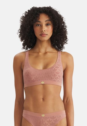 Frau mit lockigem Haar, die ein passendes Set aus rosa Sport-BH und Slip mit Adidas-Trefoil-Logo am Bund trägt, nach vorne blickend.