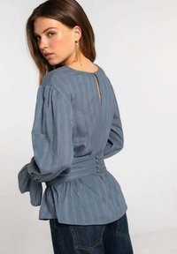 Blusa a rayas azules con mangas abullonadas, cintura fruncida y cierre de botón en la parte posterior. La tela tiene una textura suave y un corte holgado.