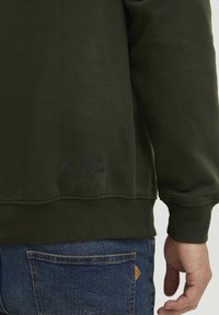 Mørkegrønn genser med ribbet kant og mansjetter. Svart "A.R.C." logo detalj på forsiden. Denim jeans delvis synlige.