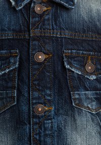 Denim jacket with dark blue wash, copper buttons, two chest pockets, and visible yellow stitching. Features distressed texture on edges.