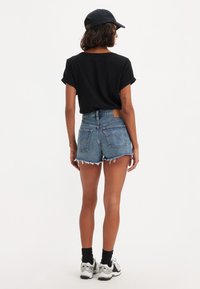 Top negro de manga corta, pantalones cortos de mezclilla azul con bordes deshilachados y calcetines negros. Combinado con zapatillas deportivas blancas y grises y una gorra negra.