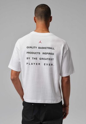 Hombre con cabello corto que lleva una camiseta blanca con texto negro y un pequeño logo rojo en la parte superior de la espalda, combinada con pantalones negros.