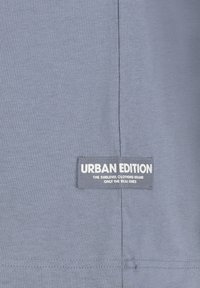 Graues Baumwoll-T-Shirt mit einem angenähten Etikett, das "URBAN EDITION" trägt und Branding-Details aufweist. Glatte Textur mit einem einfachen, lässigen Design.