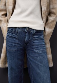 Mörkblå denimjeans med en lätt blekning, standard femfickdesign och metallknappstängning. Bärs med en ljus beige tröja och en rutiga kofta.