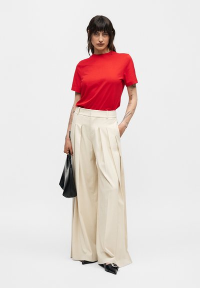 Γυναίκα που φοράει κόκκινο κοντομάνικο τοπ, ευρύ μπεζ παντελόνι με ψηλή μέση, μαύρα μυτερά παπούτσια, κρατώντας μικρή μαύρη τσάντα.