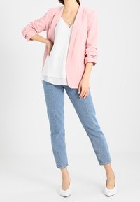 Vaaleanpunainen bleiseri käännetyillä hihoilla, valkoinen paita, jossa on ristikkäinen kaula-aukko, vaaleansiniset farkut ja valkoiset korkokengät.