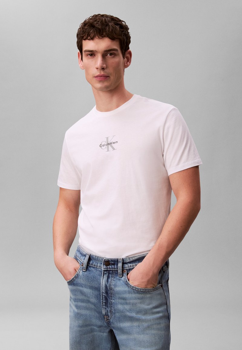 Jeune homme debout devant un fond uni, portant un t-shirt blanc avec logo Calvin Klein et un jean bleu clair, les mains dans les poches.