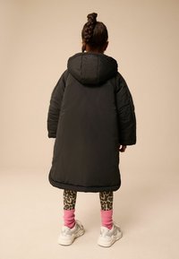 Cappotto nero imbottito con cappuccio, design oversize e texture trapuntata. Leggings trasparenti con stampa leopardata e calzini rosa a coste, abbinati a sneakers bianche.