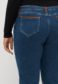 Dark blue denim jeans with a smooth texture, featuring two back pockets with brown horizontal accents and a mid-rise waist.