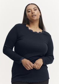 Langærmet sort top med bølgende blondedetaljer ved halsen og manchetterne, med en ribbet tekstur og lige kant. Glat stof.