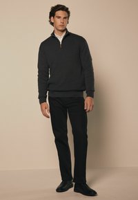 Tamno sivi zip-up pulover s visokim ovratnikom i rebrastim rubom, uparen s crnim hlačama ravnog kroja i crnim cipelama, na neutralnoj pozadini.