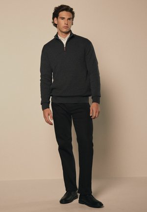 Dunkelgrauer Zip-Up-Pullover mit hohem Kragen und geripptem Saum, kombiniert mit schwarzen schmal geschnittenen Hosen und schwarzen Schuhen, vor einem neutralen Hintergrund.