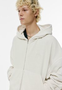 Helles beige Hoodie mit einem Frontreißverschluss, oversized Design, weicher Textur und gesticktem Logo auf der Brust. Verfügt über eine große Kordelzugkapuze.
