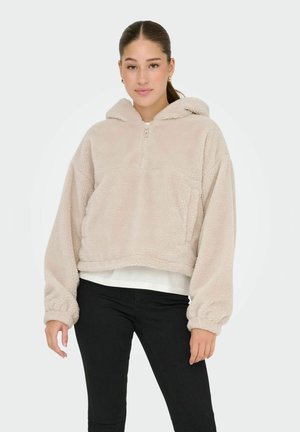 Beige fleecehoodie med halvmeterdragkedja, stor framficka och elastiska ärmslut. Bärs över en vit skjorta och matchas med svarta byxor.