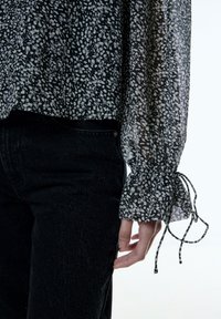 Zwart-wit gebloemd blouse van doorzichtig materiaal, met pofmouwtjes en strikdetails bij de manchetten, gecombineerd met zwarte jeans.