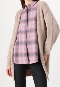 Długi, beżowy kardigan wykonany z miękkiego, teksturowanego materiału, nałożony na różowo-szarą koszulę w kratę z zapięciem na guziki. Poniżej widoczne czarne spodnie.