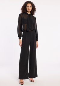 Zwarte doorzichtige blouse met lange mouwen en een strikdetail rond de nek, gecombineerd met hooggewaade zwarte wijde broek. Heeft een gladde stof en een ontspannen pasvorm.