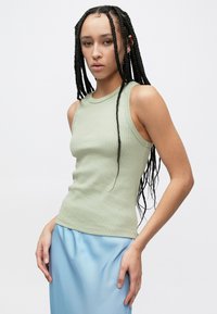 Débardeur vert clair côtelé avec encolure ronde et silhouette ajustée, associé à une jupe fluide bleu clair. Le modèle pose avec des cheveux tressés.