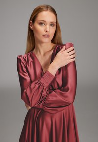 Rubinrotes Satinkleid mit tiefem V-Ausschnitt, langen Puffärmeln und gerafften Manschetten. Glatte Textur, tailliertes Oberteil und fließender Rock.