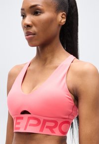 Ροζ αθλητικό σουτιέν από λείο ύφασμα, με V-λαιμόκοψη, λεπτομέρεια με άνοιγμα και φαρδιά ταινία με την ένδειξη "PRO" σε αντίθετο χρώμα.