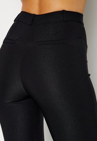 Pantaloni neri a vita alta con una texture lucida, dotati di due tasche posteriori e un design aderente che mette in risalto le curve.