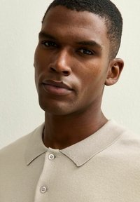 Beige poloshirt met een kraag, voorzien van twee knoopsluitingen en een fijn gebreide textuur. Model heeft kort, nauwgezet geknipt haar.