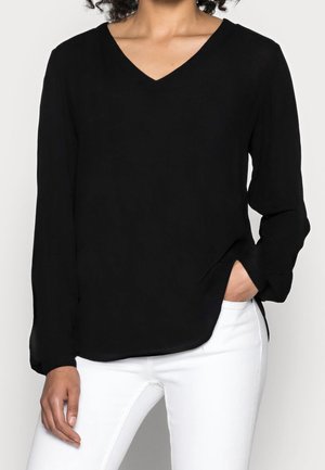 Blusa nera a maniche lunghe con scollo a V in un tessuto leggero, dal taglio rilassato, con spacchi laterali e una consistenza morbida. Abbinata a pantaloni bianchi.