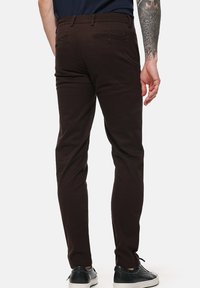 Brązowe spodnie bawełniane o dopasowanym kroju, z tylną kieszenią na zamek błyskawiczny i subtelnymi detalami w postaci przeszyć. Noszone z czarnymi sneakersami.