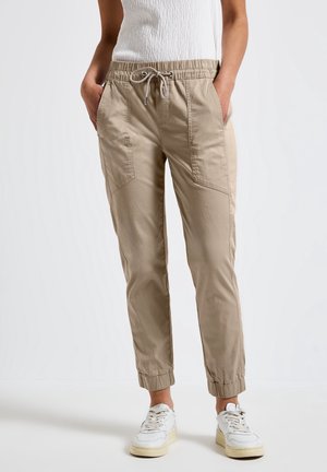 Beige broek met trekkoord en elastische boorden, gedragen met een witte mouwloze top en witte sneakers, persoon staat met handen in de zakken.
