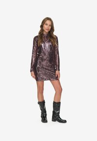 Schimmerndes, langärmliges, figurbetontes Kleid aus dunklem lila Metallic-Stoff mit vertikalen Streifen. Kombiniert mit knielangen, schwarzen, glänzenden Stiefeln.