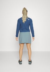 Žena s dlouhými zapletenými vlasy stojí čelem ke zdi, oblečená v modrém svetru, tyrkysové sukni a černých sportovních botách s tyrkysovými akcenty.