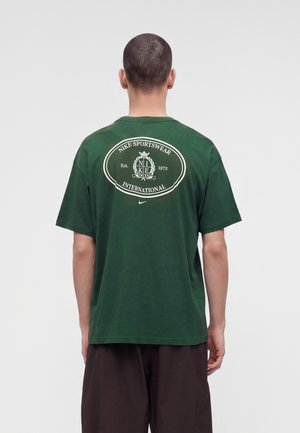 Hombre con el pelo corto que lleva una camiseta verde de Nike Sportswear International, mostrado de espaldas sobre un fondo gris liso.