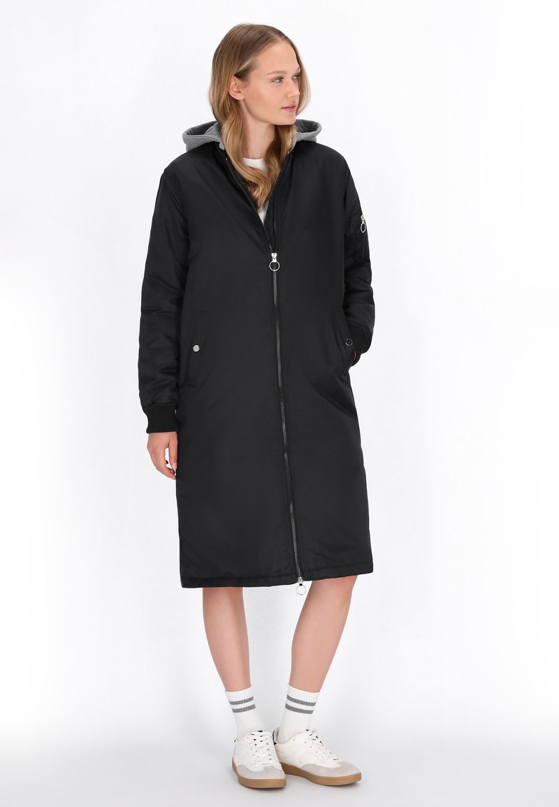 Abrigo largo negro de nailon con capucha gris, cierre con cremallera, bolsillos laterales y puños acanalados, combinado con zapatillas blancas y calcetines a rayas.