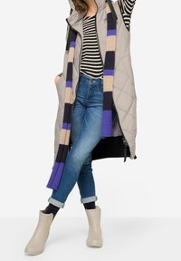 Kurtka pikowana w kolorze szarym, pasiasty top, niebieskie dżinsy, beżowe gumowe kalosze oraz dziergany szalik w fioletowe, czarne i beżowe paski.