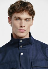 Granatowa kurtka z wysokim kołnierzem, srebrnymi zatrzaskami i przednimi kieszeniami. Gładka tekstura, dopasowany krój i brak widocznych wzorów.