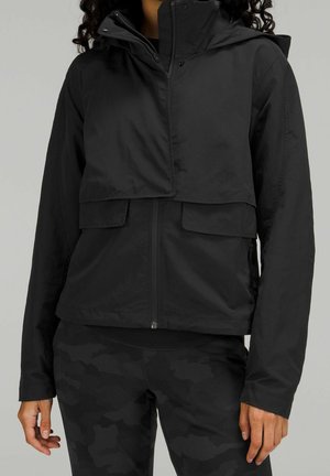 Frau trägt eine schwarze Reißverschlussjacke mit Klappentaschen und schwarz-kamouflagegemusterte Leggings vor einem einfarbigen grauen Hintergrund.