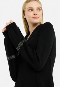 Maglietta nera a maniche lunghe con cappuccio e maniche a sbuffo, con un dettaglio del logo argentato sul petto. Tessuto morbido con una vestibilità comoda.