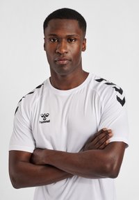 T-shirt de sport blanc avec des rayures noires sur les épaules, arborant un petit logo sur la poitrine. Texture lisse, manches courtes, col rond.