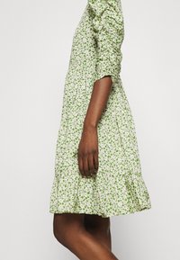 Green floral dress with white daisies, short puff sleeves, gathered waist, and ruffled hem. Made from lightweight material.