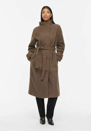 Femme portant un long manteau marron ceinturé avec un col haut, pantalon noir et bottines noires à talons, debout devant un fond blanc.