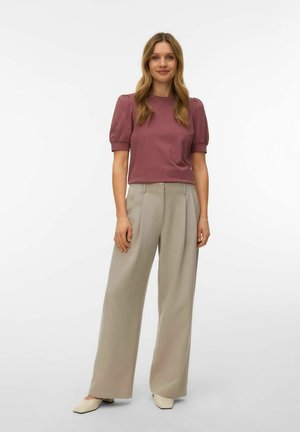 Woman standing wearing a mauve puff-sleeve top, beige wide-leg trousers, and cream square-toe shoes against a plain white background.