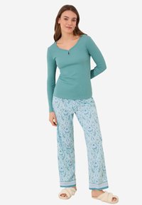 Een teal lange mouw top met een keyhole-halslijn, gecombineerd met lichtblauwe bloemenpatroon broek en crèmekleurige slippers.