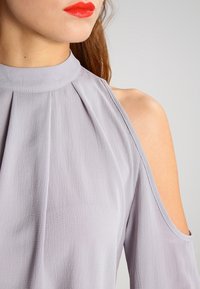 Blouse lavande claire avec un col montant, des détails plissés et un design à épaules dénudées. Tissu texturé avec une apparence douce et fluide.