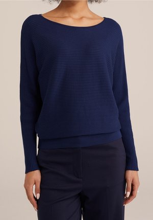 Frau trägt einen dunkelblauen, gerippten Pullover mit langen Ärmeln und Boot-Ausschnitt sowie passende dunkelblaue Hose vor neutralem Hintergrund.